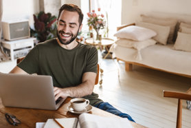 A smiling man sits at a wooden table using a laptop, with a cup of coffee, notebook, and eyeglasses nearby in a bright, cozy room with a bed and plants in the background.