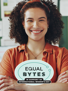 A woman with curly hair and an orange shirt smiles confidently with folded arms; a badge reads EQUAL BYTES in support of International Womens Day in front of her. Office background with charts and framed art.