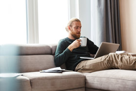 A man with a beard and tied-back hair sits on a beige couch, holding a white mug and looking at a laptop. Papers and a pen are on the sofa beside him. Large windows and gray curtains are in the background.