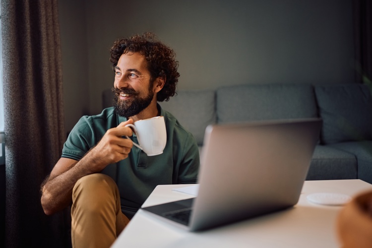 A man with curly hair and a beard sits at a table with a laptop, smiling and holding a white mug. He faces slightly away from the screen, in a cozy, well-lit room with a couch in the background.