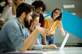 A group of young adults gathered around a laptop in an office setting, collaborating and discussing something on the screen, with one woman gesturing and others attentively engaged.