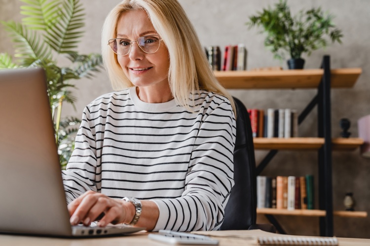 A woman with long blonde hair and glasses, wearing a striped shirt, smiles while working on a laptop at a desk. Shelves with books and plants are in the background.