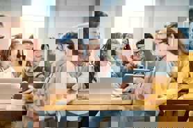 A group of young adults, apparently students, sitting around a laptop together while a lady teaches them cyber security principles by showing them something on an digital tablet.