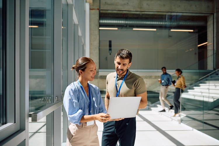 Two colleagues stand by large windows in a modern office, smiling and looking at a laptop together. In the background, two other people walk down the stairs while conversing. Bright, natural light fills the space.