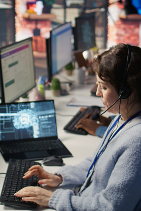 A woman wearing a headset types on a keyboard at a desk with multiple computer monitors. Another person is working in the background. The office environment appears modern and busy.