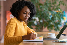 Young professional taking notes with a pencil and notepad, while sat at a laptop.