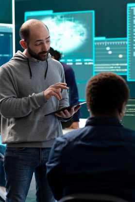 A man in a gray hoodie holds a tablet and discusses something with a colleague, who is seen from behind, in a high-tech office with large digital screens displaying data and graphics in the background.